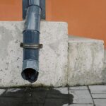 Tuyau de drainage en métal avec gouttes d'eau situé sur un mur rouge près des escaliers en béton d'un bâtiment moderne dans la rue de la ville